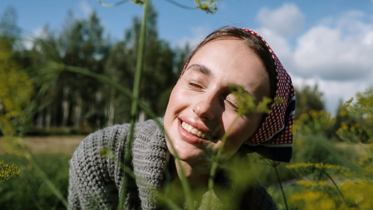 A joyful woman in a headscarf enjoying a sunny day outdoors, surrounded by greenery.